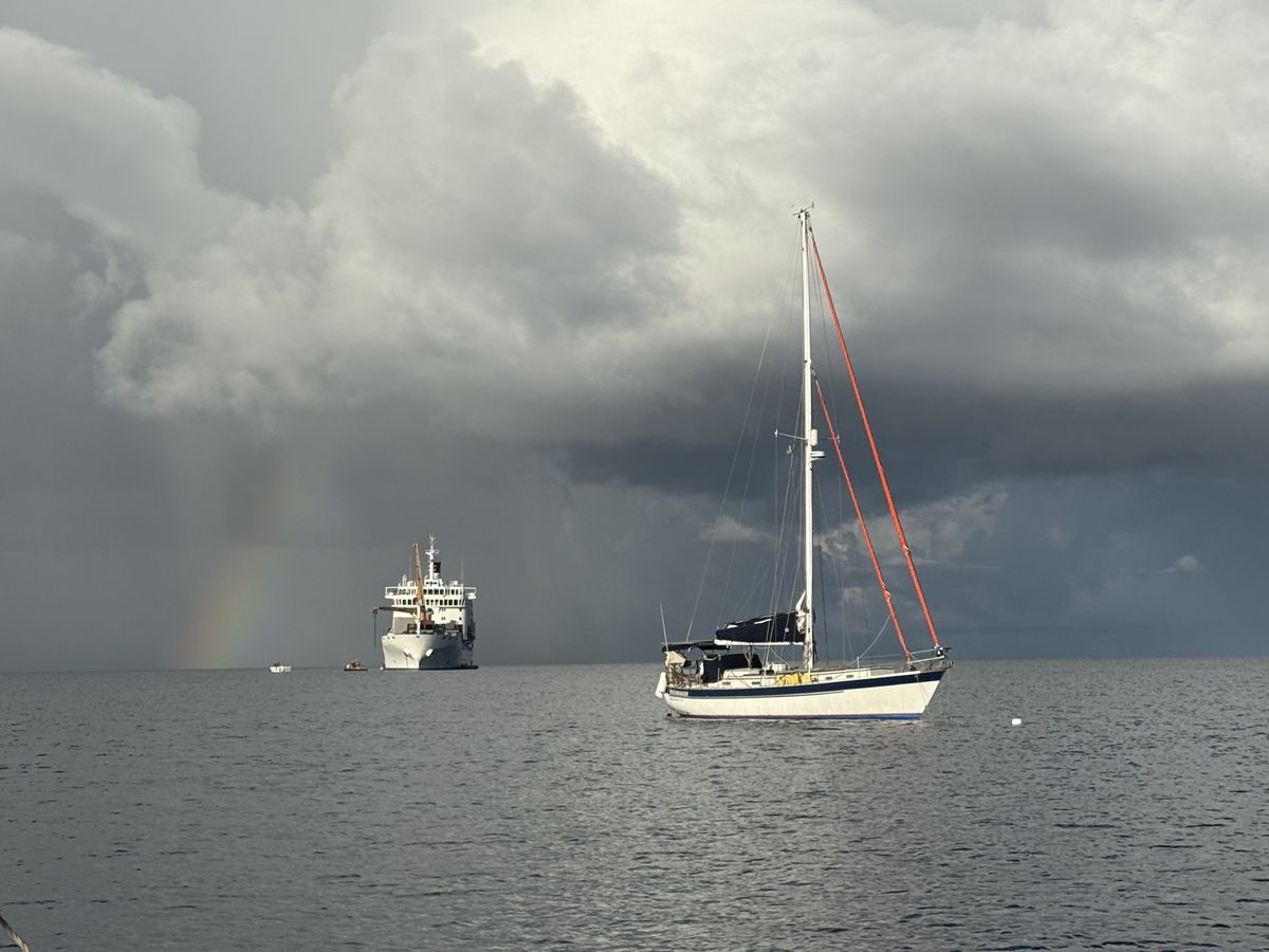 Keeldragger lying at anchor off Rangiroa, French Polynesia, with a supply ship and a rainbow under a passing squall behind her.